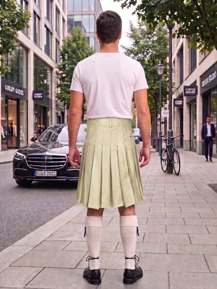 A man standing on a city sidewalk, viewed from behind, wearing a white t-shirt and a light olive green pleated kilt with white knee-high socks and black shoes.