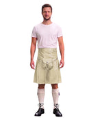 A man modeling a beige kilt with a front pouch and tassel detail, paired with a light pink t-shirt, cream knee socks, and black lace-up shoes, against a plain white background.