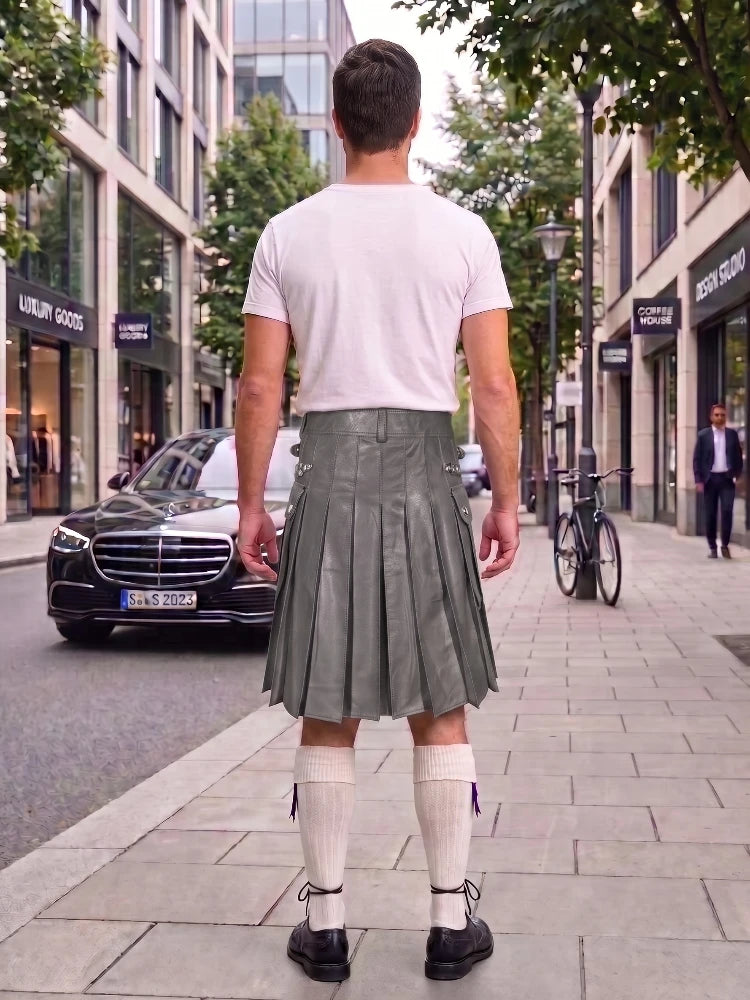 A man standing on a city sidewalk, viewed from behind, wearing a white t-shirt and a dark gray pleated leather kilt with white knee-high socks and black shoes. Storefronts, trees, and a parked black car are visible in the background.