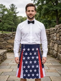 A man wearing a white dress shirt, black bow tie, and a kilt featuring an American flag pattern with white stars on a blue background and red and white side panels, standing outdoors on a stone path.