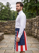 A man wearing a white dress shirt and black bow tie stands outdoors in profile, wearing a kilt with an American flag pattern featuring red and white stripes and a blue panel with white stars.