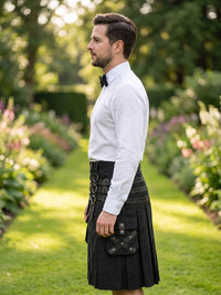 A man in a white dress shirt and black bow tie stands in profile in a sunlit garden path, wearing a black kilt with a leather belt and a black leather sporran pouch.