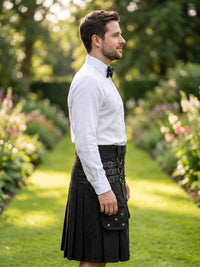 A man standing in profile in a sunlit garden path, wearing a white long-sleeve dress shirt, black bow tie, and a black kilt with a front flap pocket.