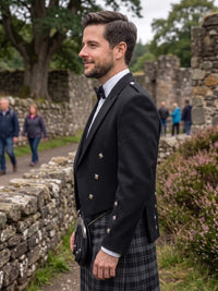 A man wearing a traditional Scottish formal outfit with a black double-breasted jacket, white shirt, black bow tie, and a gray plaid kilt, standing outdoors in front of stone ruins.