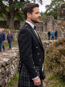 A man in profile wearing a black double-breasted jacket with silver buttons and a black-and-gray tartan kilt, standing outdoors near stone ruins and other blurred people in the background.