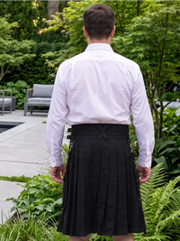 A man viewed from behind wearing a white dress shirt and a black pleated kilt in an outdoor garden setting.