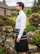 A man wearing a black kilt with gray and black striped waist panel, a white dress shirt, and a black bow tie, standing outdoors in a rural Scottish landscape with a stone cottage and green hills in the background.