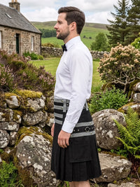 A man wearing a black kilt with gray and black striped waist panel, a white dress shirt, and a black bow tie, standing outdoors in a rural Scottish landscape with a stone cottage and green hills in the background.