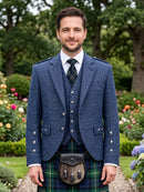 A man wearing a traditional Scottish formal outfit with a textured blue blazer, matching waistcoat, a green and blue plaid kilt, and a black sporran bag, standing in a lush garden setting.