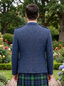 A man viewed from behind wearing a textured navy blue tweed blazer and a green and blue tartan kilt, standing in a lush garden setting.