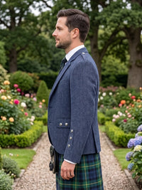 A man in profile wearing a navy blue herringbone blazer and a green tartan kilt, standing in a lush garden with flower beds and trees in the background.