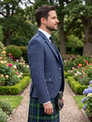 A man in profile wearing a blue tweed blazer, white dress shirt, dark tie, and a green tartan kilt with a black sporran, standing in a garden path.