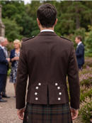 A man viewed from behind wearing a dark brown formal jacket with silver decorative buttons and a tartan kilt, standing outdoors in a garden setting with blurred guests in the background.