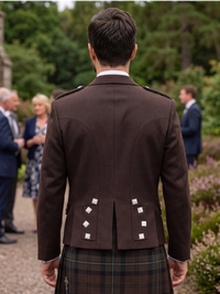A man viewed from behind wearing a dark brown formal jacket with silver decorative buttons and a tartan kilt, standing outdoors in a garden setting with blurred guests in the background.