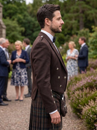 A man in a formal outdoor setting wearing a dark brown double-breasted jacket with silver buttons, a white shirt, a black bow tie, and a tartan kilt. Guests are blurred in the background among green trees and plants.