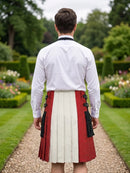 A man viewed from behind wearing a white long-sleeve shirt and a pleated kilt with vertical panels in red, off-white, and black, standing on a garden path.