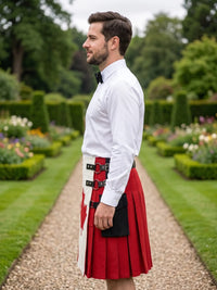 A man in a white dress shirt and black bow tie stands in profile on a garden path wearing a red and white kilt with a black belt and a black sporran pouch.