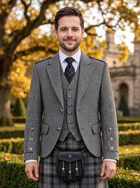 A man wearing a formal Scottish outfit with a gray herringbone blazer, matching vest and tie, a plaid kilt, and a black sporran bag, standing outdoors in front of a blurred garden and stone building.