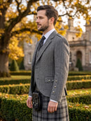 A man wearing a gray herringbone blazer, white dress shirt, black tie, and a gray plaid kilt stands outdoors in front of a stone building and autumn trees with yellow leaves.