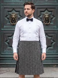 A man wearing a white pinstriped shirt, black bow tie, and a gray and white herringbone patterned kilt stands in front of a large green metal door with lion head knobs.