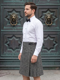 A man wearing a white long-sleeve dress shirt, black bow tie, and a gray herringbone pleated skirt, standing in front of a dark green ornate wooden door with lion head knobs.