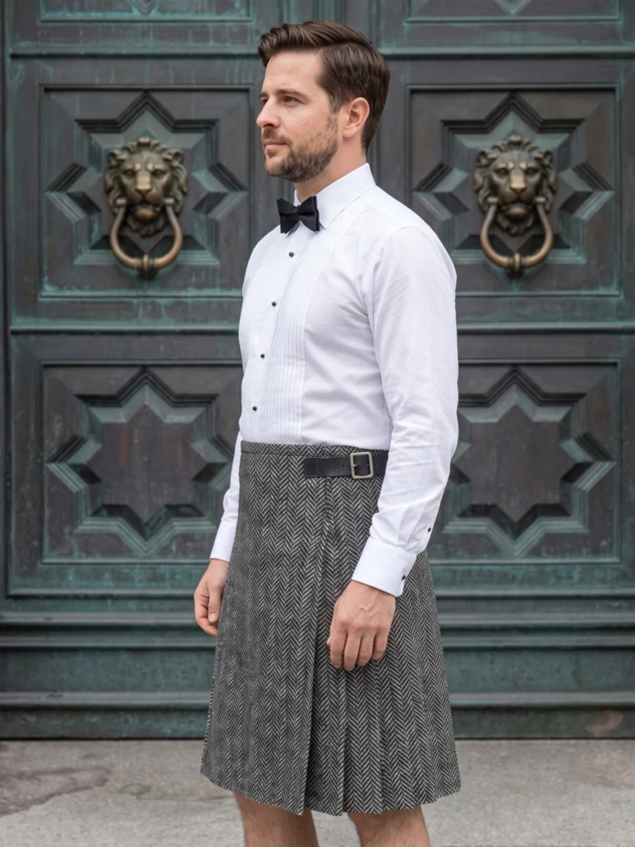 A man wearing a white long-sleeve dress shirt, black bow tie, and a gray herringbone pleated skirt, standing in front of a dark green ornate wooden door with lion head knobs.