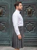 A man wearing a white long-sleeve dress shirt, black bow tie, and a gray herringbone kilt with a black belt and silver buckles, standing in front of ornate dark green wooden doors.