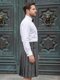 A man wearing a white long-sleeve dress shirt, black bow tie, and a gray herringbone kilt with a black belt and silver buckles, standing in front of ornate dark green wooden doors.