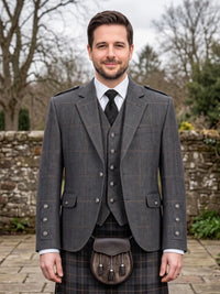 A man wearing a traditional Scottish formal outfit with a gray windowpane plaid jacket, matching waistcoat, kilt, and a brown leather sporran, standing outdoors in front of a stone wall.