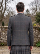 A man viewed from behind wearing a dark gray windowpane plaid blazer and a matching plaid kilt, standing outdoors in front of a stone wall and bare trees.