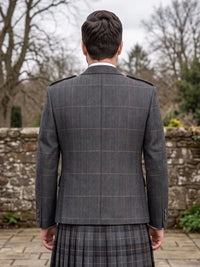 A man viewed from behind wearing a dark gray windowpane plaid blazer and a matching plaid kilt, standing outdoors in front of a stone wall and bare trees.