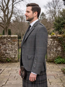 A man in profile wearing a gray windowpane plaid blazer, a dark plaid kilt, a white dress shirt, and a black tie, holding a black sporran outdoors in front of a stone wall.