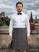 A man wearing a white pinstriped shirt, black bow tie, and a gray plaid kilt stands outdoors in front of Edinburgh's historic skyline under a cloudy sky.