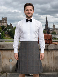A man wearing a white pinstriped shirt, black bow tie, and a gray plaid kilt stands outdoors in front of Edinburgh's historic skyline under a cloudy sky.