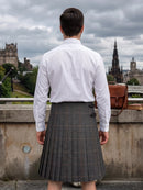A man viewed from behind wearing a white long-sleeve dress shirt and a gray plaid kilt, standing outdoors with Edinburgh's historic skyline in the background under a cloudy sky.