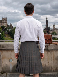 A man viewed from behind wearing a white long-sleeve dress shirt and a gray plaid kilt, standing outdoors with Edinburgh's historic skyline in the background under a cloudy sky.