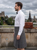 A man wearing a gray plaid kilt, white dress shirt, and black bow tie stands outdoors in Edinburgh, with a brown leather satchel bag and city skyline in the background.