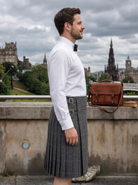 A man in profile wearing a white dress shirt, black bow tie, and gray plaid kilt, standing outdoors on a stone bridge with Edinburgh castle and spires in the background. A brown leather satchel rests on the wall beside him.
