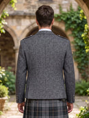 A man viewed from behind wearing a gray textured blazer with shoulder epaulets and a plaid kilt, standing in front of a blurred stone archway with green ivy.