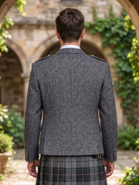 A man viewed from behind wearing a gray textured blazer with shoulder epaulets and a plaid kilt, standing in front of a blurred stone archway with green ivy.