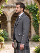 A man in profile wearing a gray tweed blazer, white dress shirt, dark tie, and a gray plaid kilt, standing outdoors in front of a stone courtyard with arches and greenery.