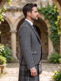 A man in profile wearing a gray tweed kilt jacket, white dress shirt, black tie, and a gray plaid kilt, standing in a stone courtyard with arches and greenery.