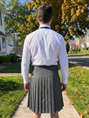 A man viewed from behind wearing a white long-sleeve button-up shirt and a dark gray pleated kilt, standing on a suburban sidewalk with trees and houses in the background.