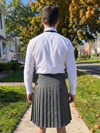 A man viewed from behind wearing a white long-sleeve button-up shirt and a dark gray pleated kilt, standing on a suburban sidewalk with trees and houses in the background.