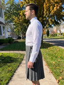 A man standing outdoors on a sidewalk wearing a white long-sleeve dress shirt, black bow tie, and a gray pleated kilt.