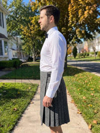 A man standing outdoors on a sidewalk wearing a white long-sleeve dress shirt, black bow tie, and a gray pleated kilt.