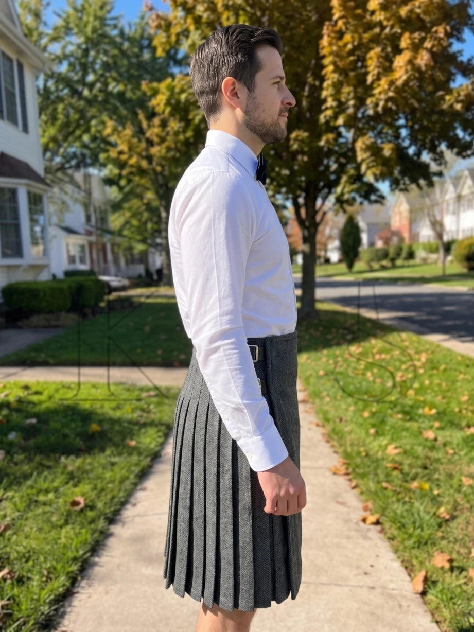 A man wearing a white long-sleeve dress shirt and a dark gray pleated kilt stands on a suburban sidewalk, shown in profile against a background of trees and houses.
