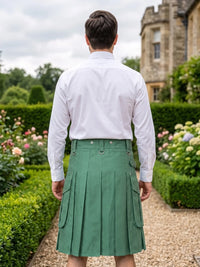 A man viewed from behind wearing a white long-sleeve dress shirt and a green pleated kilt, standing on a garden path with hedges and a stone building in the background.