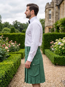 A man in a garden wearing a white long-sleeve dress shirt and a green kilt with front flap pockets, standing on a gravel path with hedges and a stone building in the background.