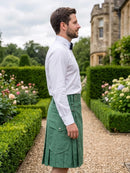 A man in a garden wearing a white long-sleeve dress shirt and a green pleated kilt, shown in profile. The background features a stone castle, manicured hedges, and flower beds.